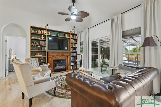 living area with a glass covered fireplace, a ceiling fan, wood finished floors, arched walkways, and vaulted ceiling