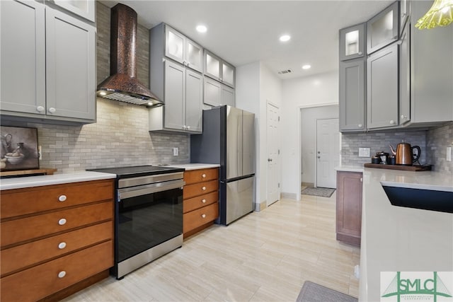 kitchen with appliances with stainless steel finishes, wall chimney exhaust hood, light countertops, decorative backsplash, and glass insert cabinets