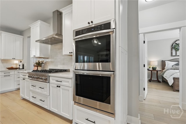 kitchen featuring appliances with stainless steel finishes, wall chimney exhaust hood, decorative backsplash, light wood-style floors, and white cabinetry
