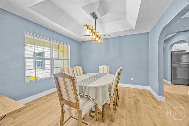dining space with arched walkways, a tray ceiling, light wood-style floors, a textured wall, and baseboards