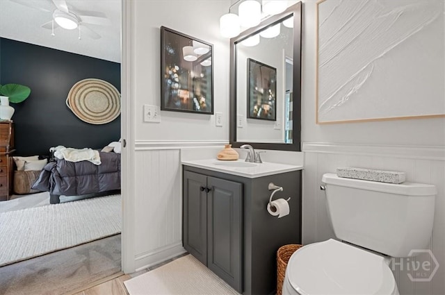 half bath with a wainscoted wall, vanity, toilet, a decorative wall, and a ceiling fan