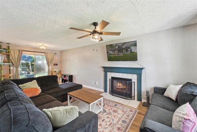living area featuring a textured ceiling, light wood-style flooring, a tiled fireplace, ceiling fan, and baseboards