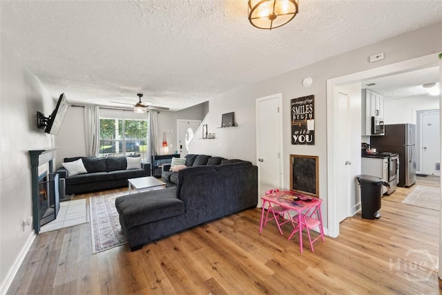 living area featuring a textured ceiling, light wood-type flooring, a fireplace with flush hearth, ceiling fan, and baseboards