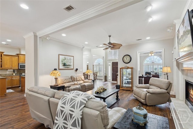 living area with a tiled fireplace, a ceiling fan, dark wood-style floors, ornamental molding, and recessed lighting