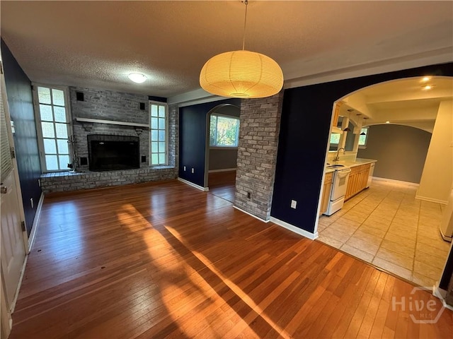 unfurnished living room with arched walkways, light wood-style flooring, baseboards, a brick fireplace, and a textured ceiling