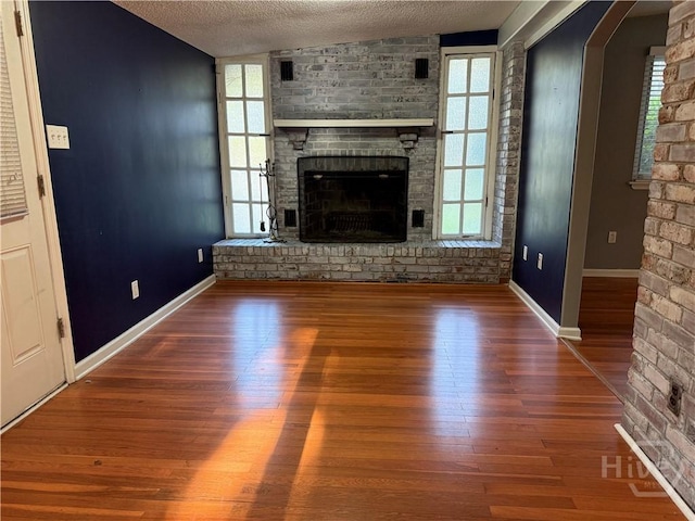unfurnished living room with a textured ceiling, a brick fireplace, wood finished floors, baseboards, and healthy amount of natural light