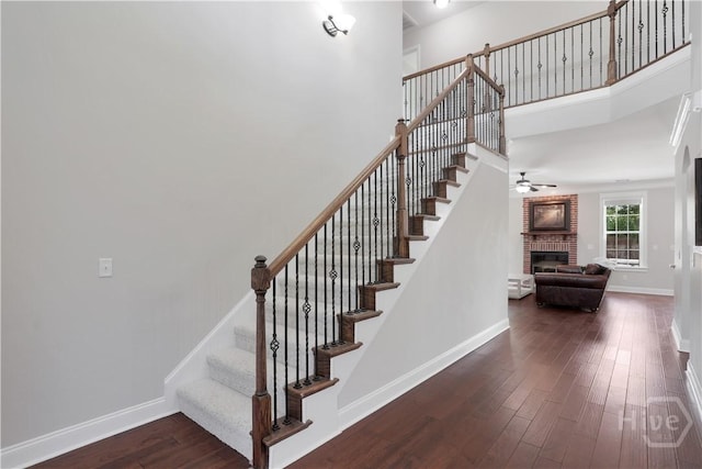 staircase with a ceiling fan, a brick fireplace, wood finished floors, baseboards, and a towering ceiling