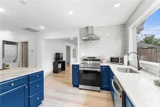 911 Hamilton Ct, Savannah, GA 31401 - kitchen featuring appliances with stainless steel finishes, blue cabinetry, wall chimney range hood, stacked washer / drying machine, and light stone countertops