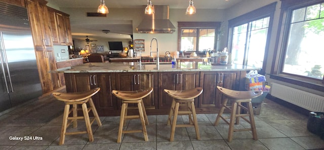 bar with wall chimney range hood, radiator, a wealth of natural light, stainless steel refrigerator, and ceiling fan