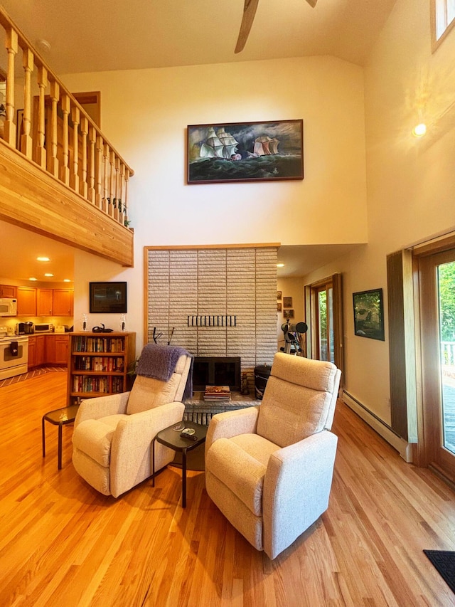 living area with a high ceiling, a baseboard heating unit, light wood-style floors, a brick fireplace, and a ceiling fan