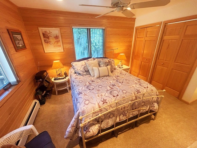 carpeted bedroom with a baseboard heating unit, wood walls, multiple closets, and ceiling fan