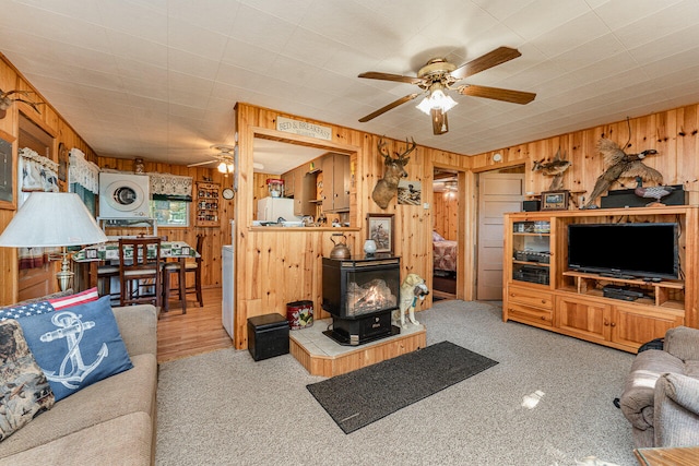 living room featuring ceiling fan, a wood stove, light carpet, and wood walls