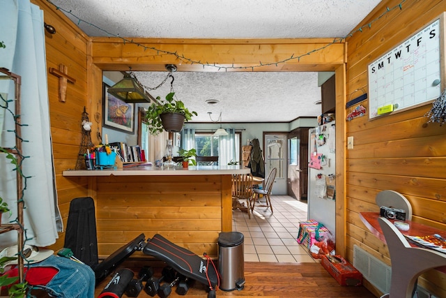 kitchen with a textured ceiling, wooden walls, hanging light fixtures, stainless steel fridge, and light hardwood / wood-style floors