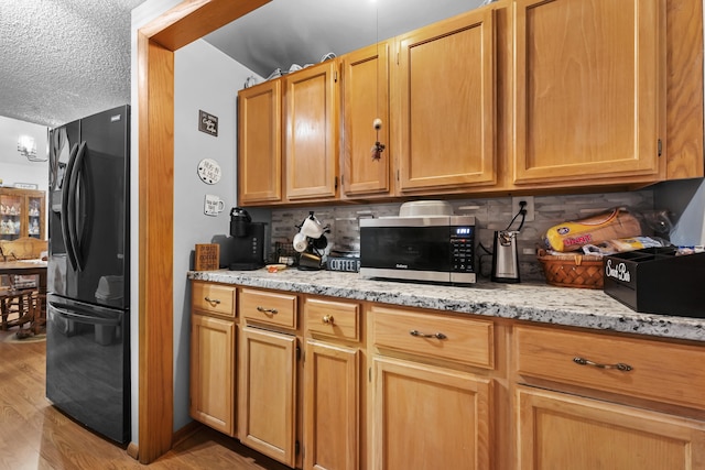 kitchen featuring light stone counters, light hardwood / wood-style floors, a textured ceiling, backsplash, and black refrigerator