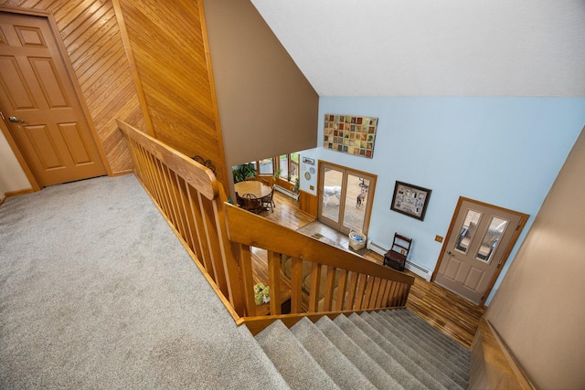 stairway featuring baseboard heating, high vaulted ceiling, wooden walls, and carpet