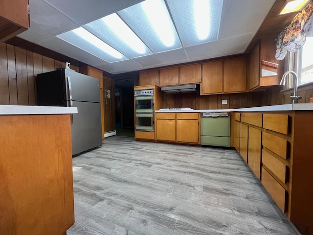 kitchen with double oven, range hood, dishwasher, stainless steel refrigerator, and wood walls