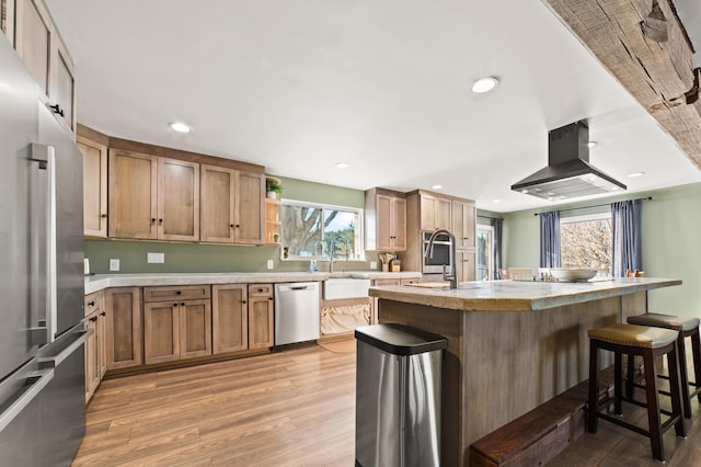 kitchen with a kitchen bar, appliances with stainless steel finishes, island range hood, a kitchen island with sink, and light hardwood / wood-style flooring