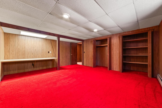 basement featuring built in shelves, wood walls, a drop ceiling, and a baseboard radiator