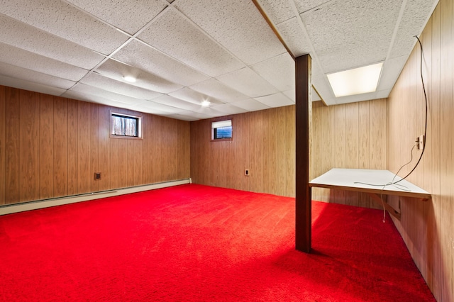 basement with carpet flooring, a drop ceiling, wooden walls, and a baseboard heating unit