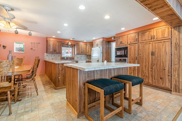 kitchen featuring a kitchen breakfast bar, black microwave, decorative backsplash, tile countertops, and white fridge