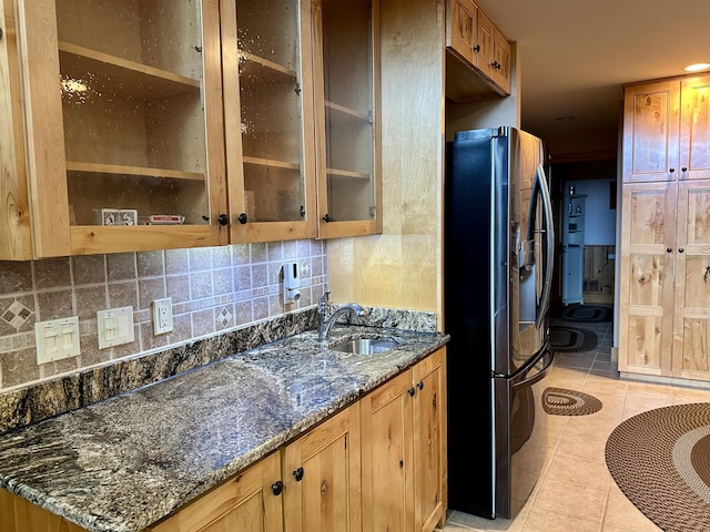 kitchen featuring light tile patterned floors, stainless steel fridge, dark stone counters, a sink, and backsplash