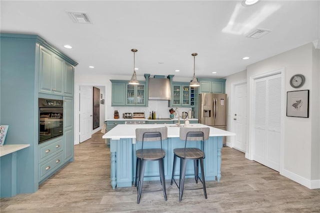 kitchen with visible vents, black oven, light countertops, wall chimney exhaust hood, and stainless steel fridge