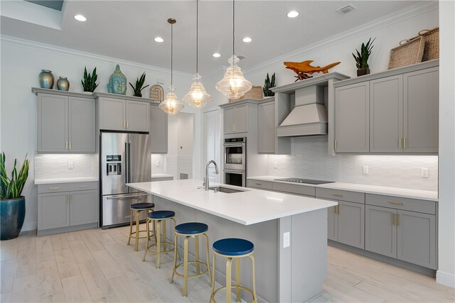 kitchen with a kitchen bar, a center island with sink, stainless steel appliances, gray cabinetry, and custom range hood