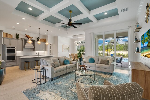 living room with coffered ceiling, beamed ceiling, light wood-type flooring, ceiling fan with notable chandelier, and sink