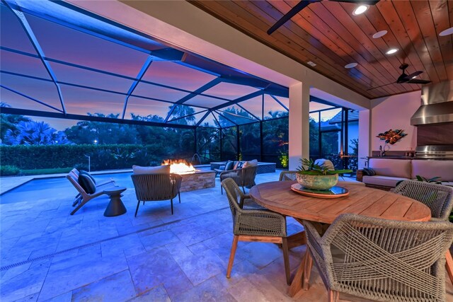 patio terrace at dusk with area for grilling, ceiling fan, glass enclosure, and an outdoor fire pit