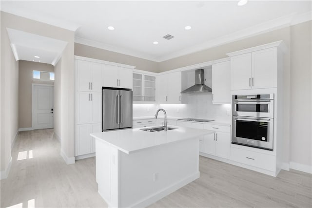 kitchen featuring a center island with sink, wall chimney range hood, sink, appliances with stainless steel finishes, and white cabinetry