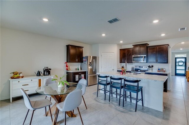 kitchen featuring appliances with stainless steel finishes, dark brown cabinets, a kitchen island with sink, and light tile patterned flooring