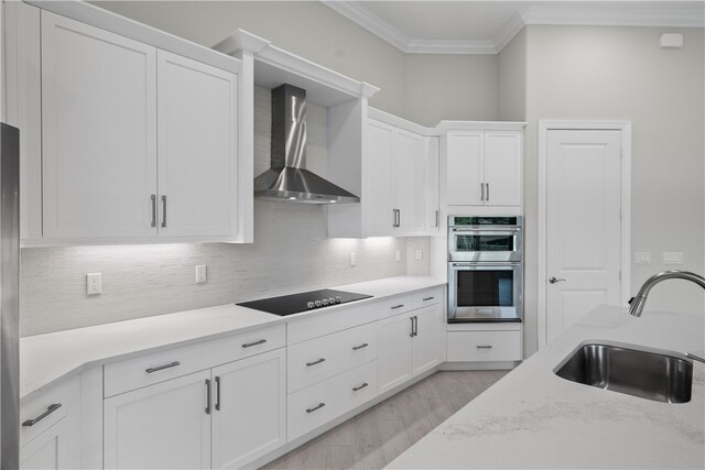 kitchen with black electric stovetop, white cabinetry, wall chimney range hood, sink, and double oven