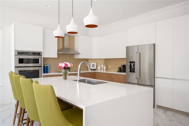 kitchen with appliances with stainless steel finishes, backsplash, wall chimney range hood, white cabinetry, and an island with sink