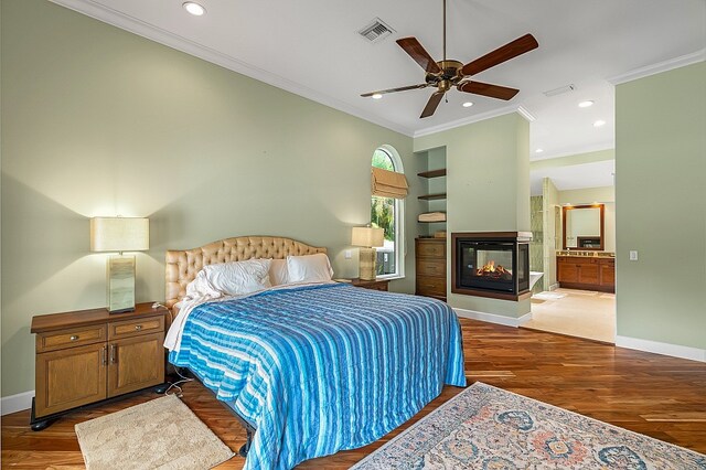 bedroom featuring wood-type flooring, crown molding, ensuite bath, a multi sided fireplace, and ceiling fan