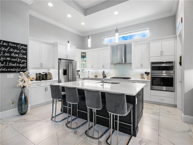 kitchen featuring stainless steel appliances, wall chimney range hood, sink, white cabinets, and a kitchen island with sink