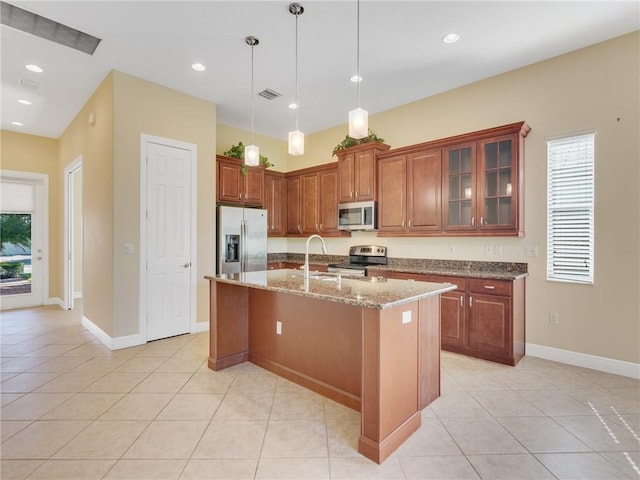 kitchen featuring light tile patterned floors, hanging light fixtures, stainless steel appliances, light stone counters, and an island with sink