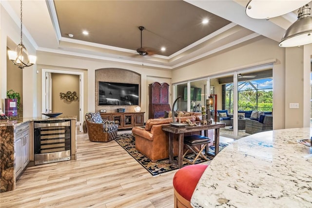 living room featuring ceiling fan with notable chandelier, a raised ceiling, crown molding, light hardwood / wood-style floors, and beverage cooler