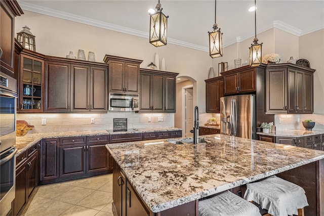 kitchen featuring crown molding, pendant lighting, a kitchen island with sink, dark brown cabinets, and appliances with stainless steel finishes