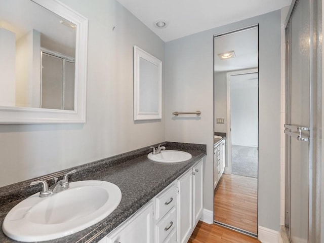 full bath with a shower with shower door, double vanity, a sink, and wood finished floors