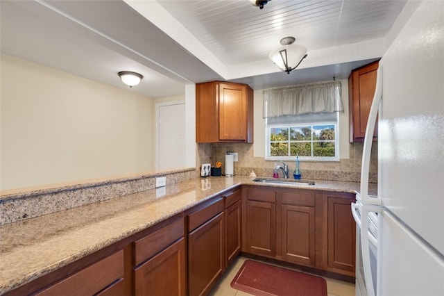 kitchen with backsplash, light stone counters, freestanding refrigerator, brown cabinetry, and a sink