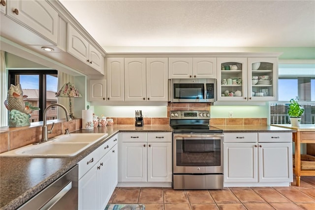 kitchen featuring stainless steel appliances, a sink, white cabinets, dark countertops, and glass insert cabinets