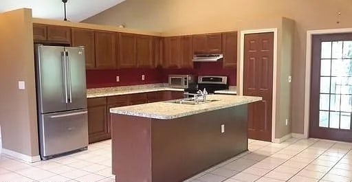 kitchen featuring stainless steel fridge, light stone counters, a kitchen island with sink, light tile patterned floors, and range