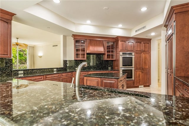 kitchen featuring premium range hood, visible vents, backsplash, stainless steel double oven, and glass insert cabinets