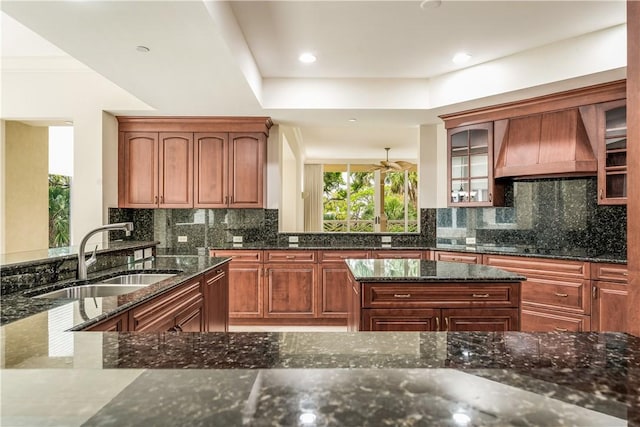 kitchen with a sink, dark stone countertops, backsplash, glass insert cabinets, and custom exhaust hood