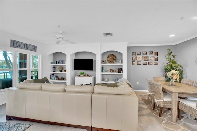 living area with built in features, visible vents, light wood-style flooring, ornamental molding, and ceiling fan