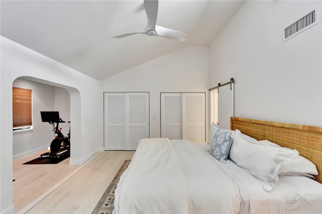 bedroom featuring ceiling fan, high vaulted ceiling, multiple closets, light hardwood / wood-style flooring, and a barn door