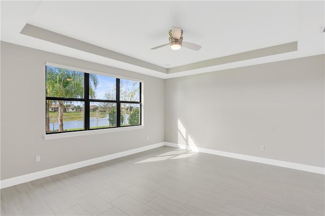 unfurnished room with a ceiling fan, a raised ceiling, a water view, and baseboards