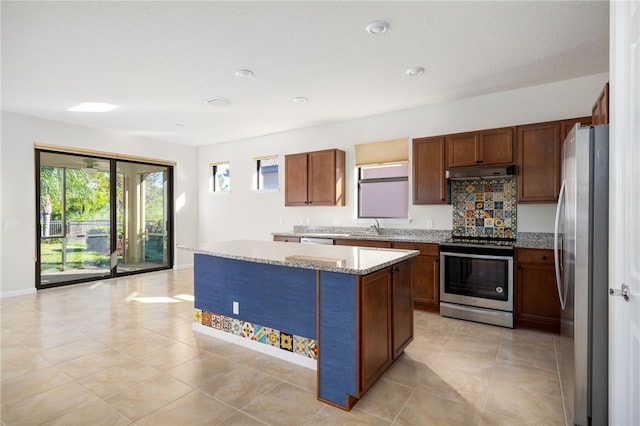 kitchen with a kitchen island, appliances with stainless steel finishes, brown cabinets, light stone counters, and under cabinet range hood