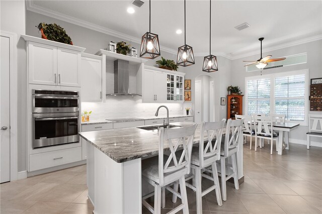 kitchen with white cabinets, wall chimney exhaust hood, sink, stainless steel double oven, and a kitchen island with sink