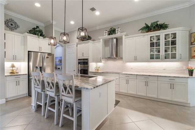 kitchen featuring a center island with sink, white cabinets, wall chimney range hood, double oven, and decorative light fixtures